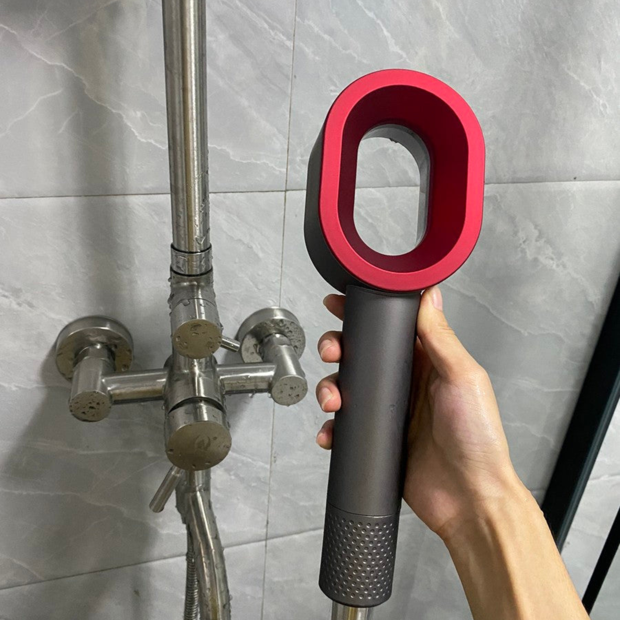 Hand holding a red and black handheld shower head against a tiled bathroom wall.